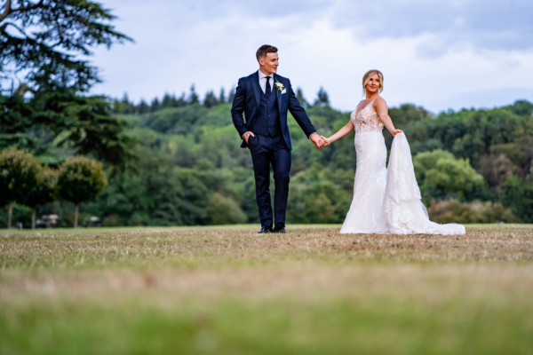 Wedding couple stand together on lawns whilst bride holds the train of her dress